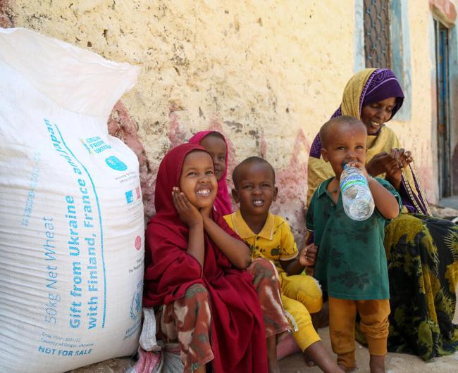 A group of children sit outside a house next to a WFP branded bag of wheat.